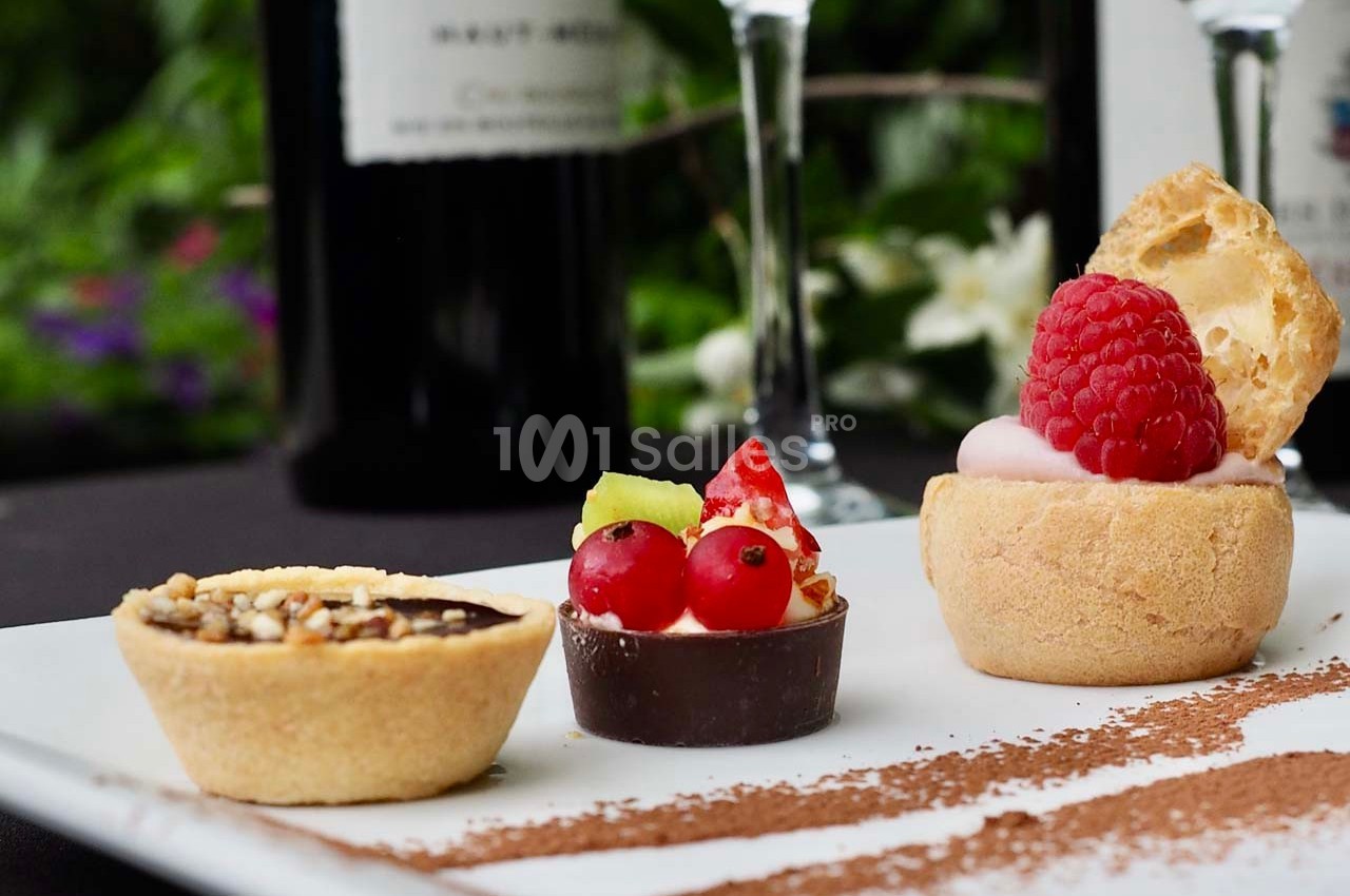 Assortiment de desserts miniatures sur une assiette blanche, décorée de cacao en poudre, avec un fond flou de bouteilles de…
