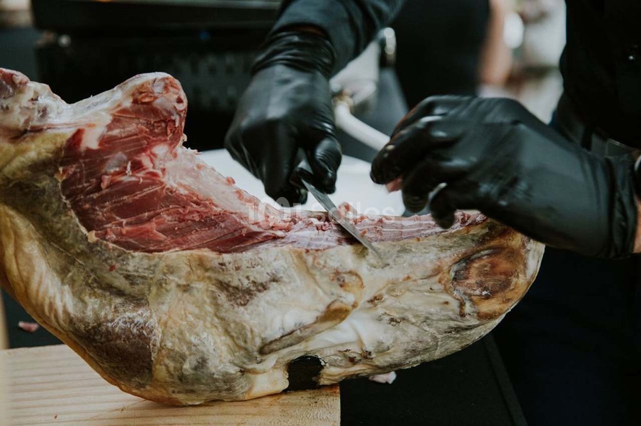 Une personne découpe un jambon sec sur un support en bois avec un couteau, portant des gants noirs.
