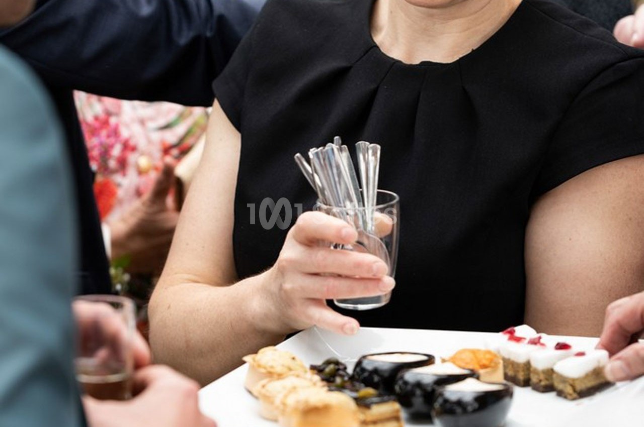 Une personne en tenue noire tient un verre rempli de cuillères, entourée de convives prenant des amuse-bouches sur un…