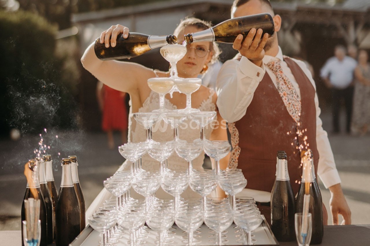 Un couple verse du champagne dans une pyramide de coupes en extérieur, entouré de bouteilles et d'invités en arrière-plan.