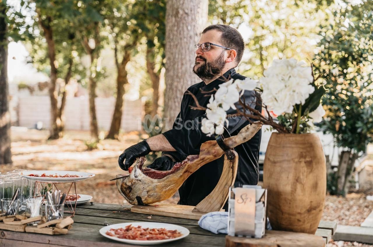Un homme découpe un jambon cru sur une table en bois, entouré d'assiettes et de couverts, dans un cadre extérieur boisé.