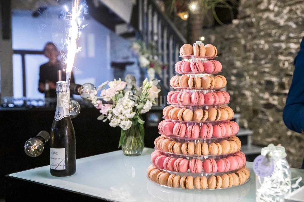 Tour de macarons roses et beiges sur une table avec une bouteille de champagne et un bouquet de fleurs en arrière-plan.