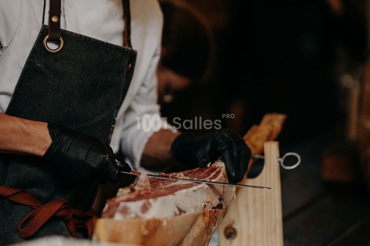Un cuisinier en tablier tranche délicatement un jambon posé sur un support en bois.