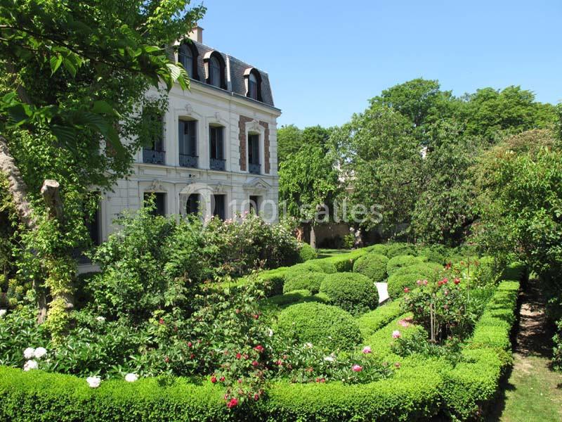 Jardin paysager avec buissons taillés, rosiers et façade d'une maison blanche à plusieurs étages sous un ciel dégagé.