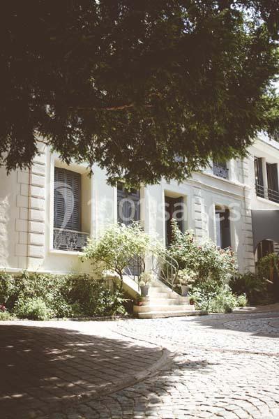 Façade d'une maison blanche avec volets bleus, entourée de végétation et bordée d'une allée pavée.