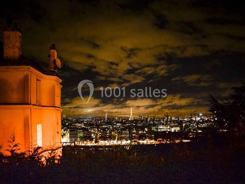 Vue nocturne d'une ville illuminée avec une maison éclairée au premier plan et une tour visible à l'horizon.