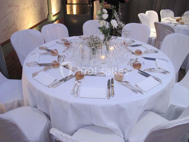 Table ronde dressée pour un repas, avec nappes blanches, couverts, verres, serviettes, petits pains et centre floral.