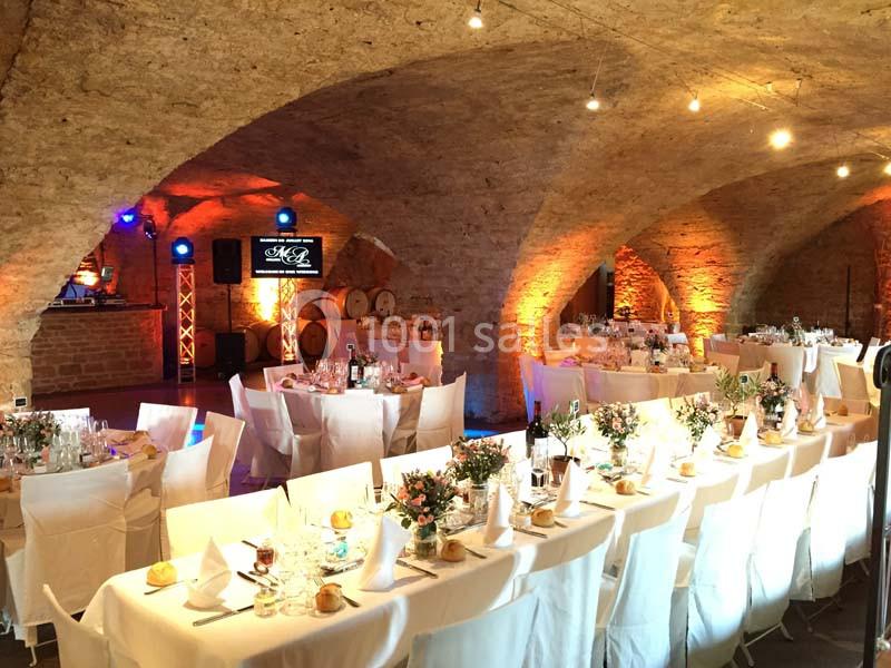 Salle voûtée en pierre décorée pour un événement, avec tables dressées, chaises blanches et éclairage chaleureux.