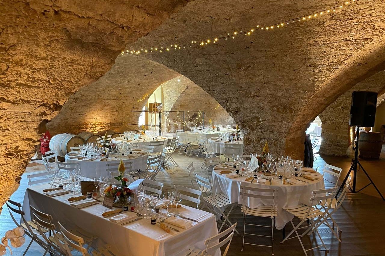 Salle voûtée en pierre aménagée pour un événement, avec tables rondes et rectangulaires décorées, éclairage chaleureux.