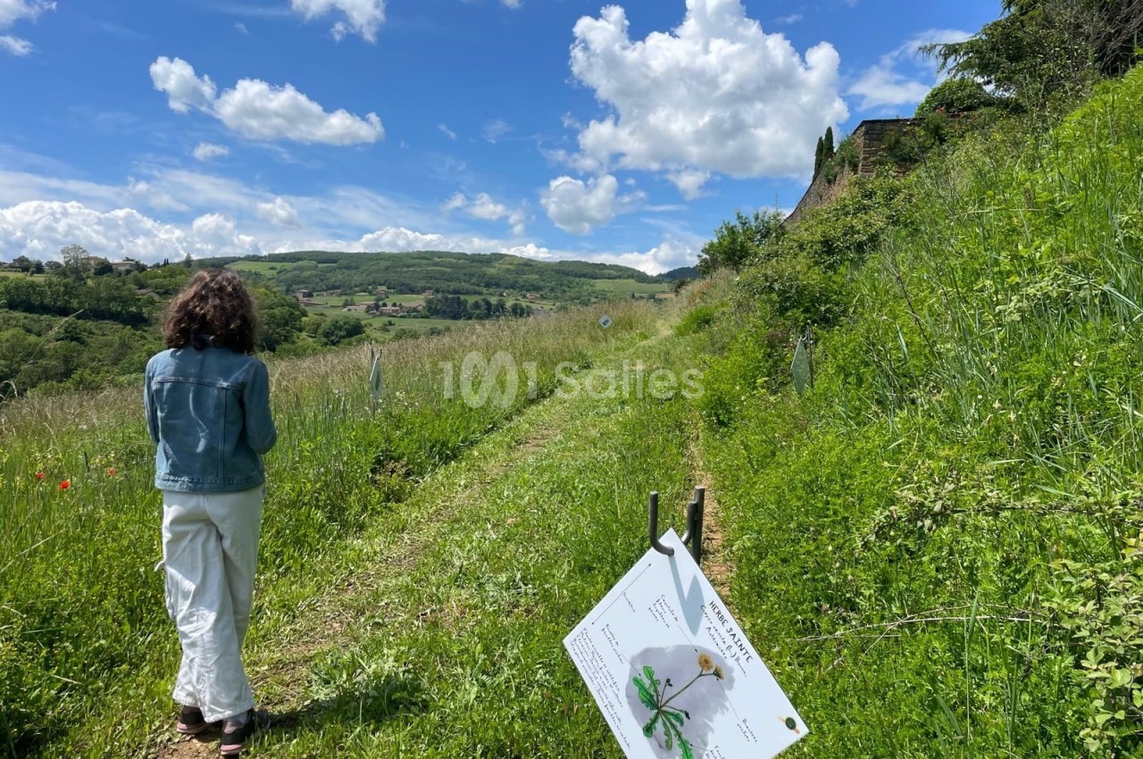 Une femme marche sur un sentier bordé de verdure et de panneaux explicatifs dans un paysage vallonné.