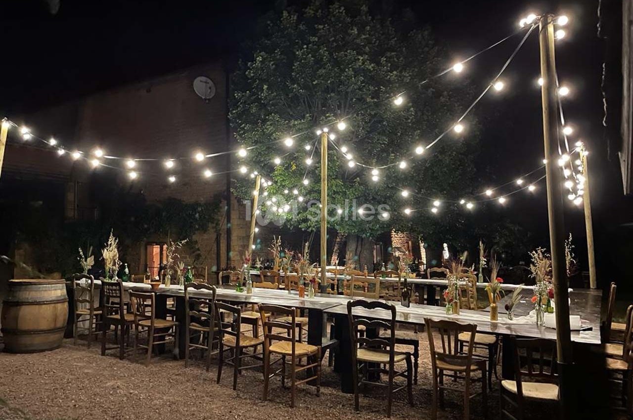 Tables en bois décorées de fleurs et chaises dépareillées sous des guirlandes lumineuses dans une cour extérieure la nuit.