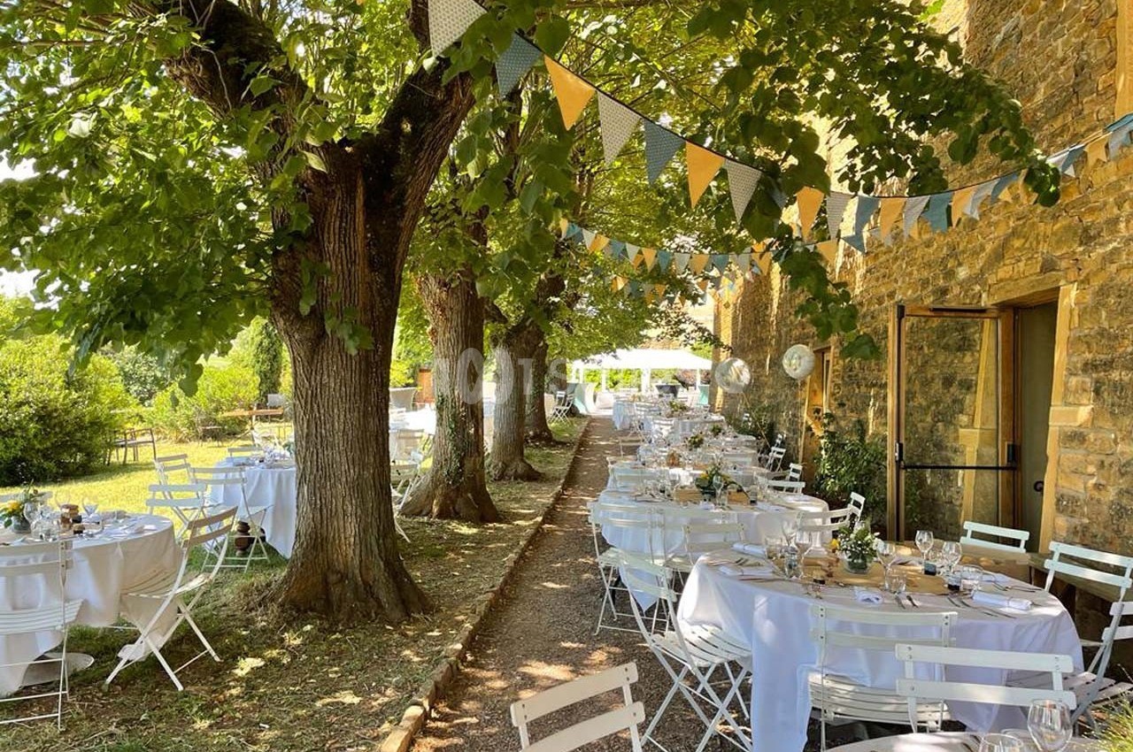 Tables décorées pour un repas en plein air sous des arbres, avec guirlandes suspendues et bâtiment en pierre en arrière-plan.