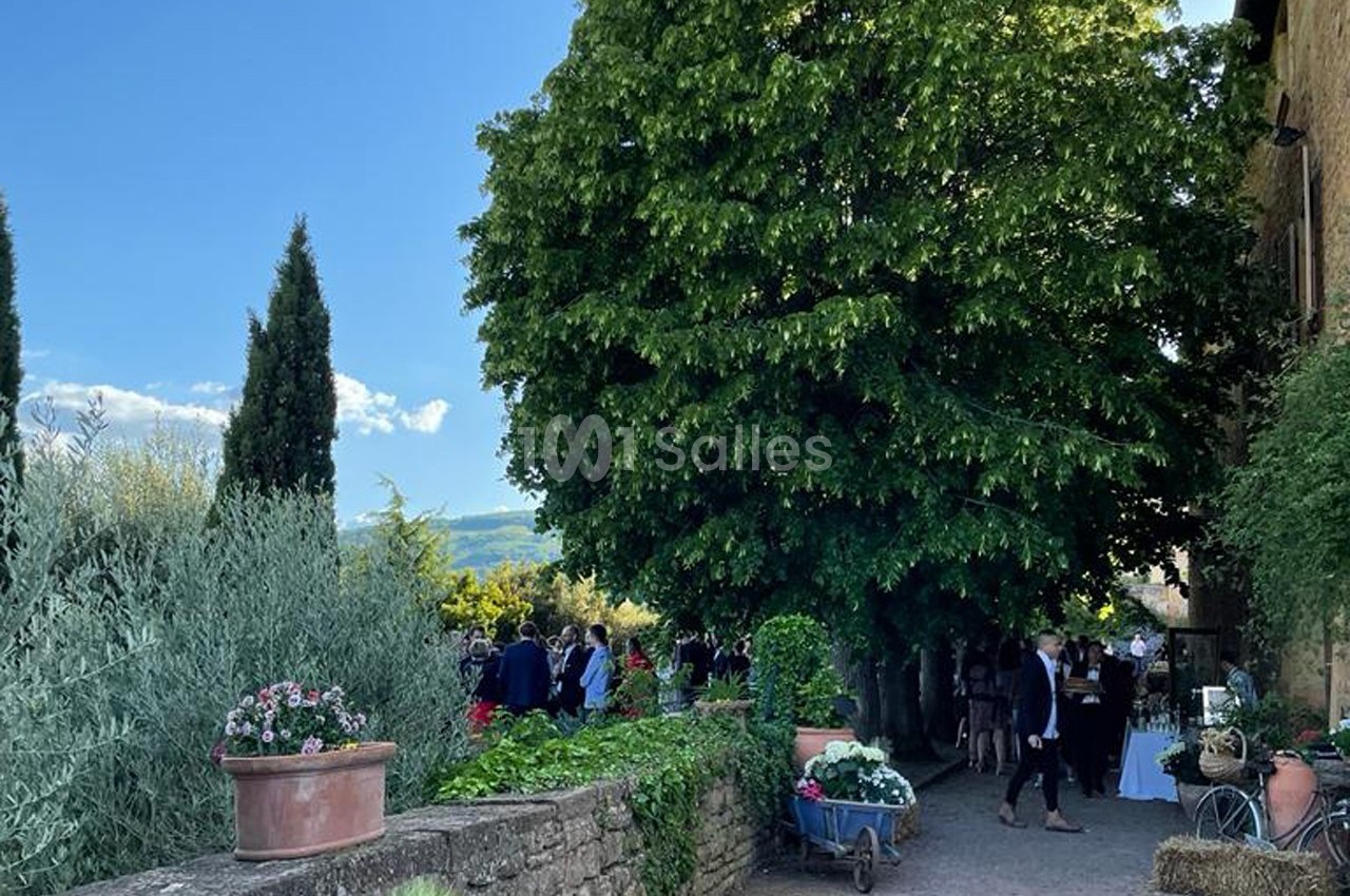 Un chemin bordé de végétation mène à un rassemblement sous un grand arbre, avec des pots de fleurs et une charrette…