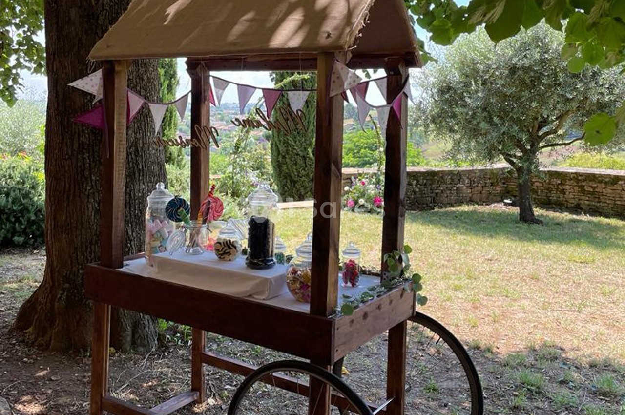 Chariot en bois décoré avec des bonbonnières et des guirlandes, installé dans un jardin ombragé.