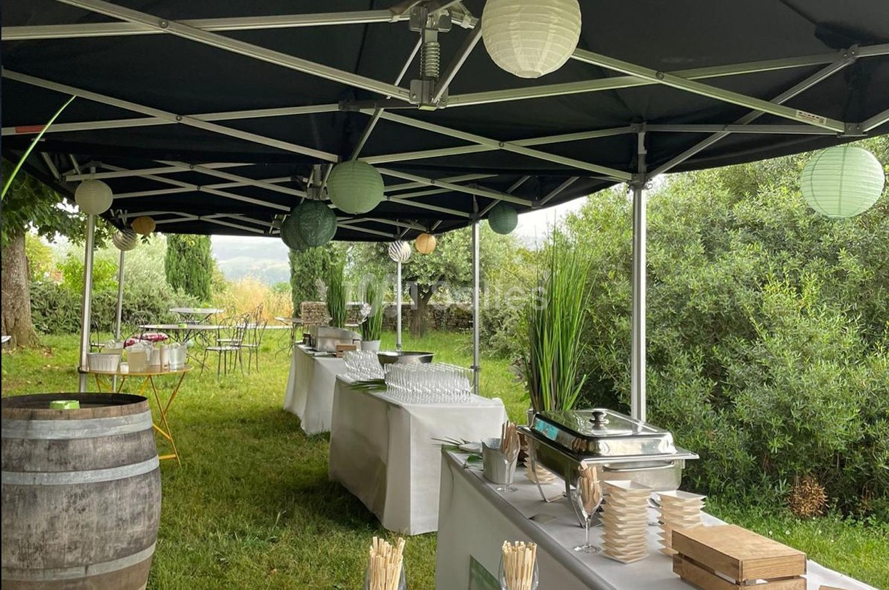 Buffet en extérieur sous des tonnelles noires, avec vaisselle, plats et décorations lumineuses dans un cadre verdoyant.