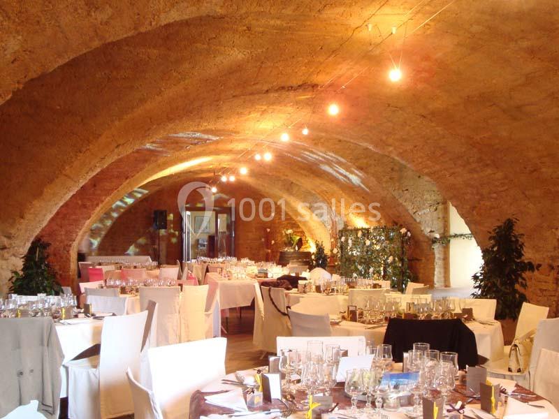 Salle voûtée en pierre aménagée pour un événement, avec tables dressées, nappes blanches et éclairage chaleureux.
