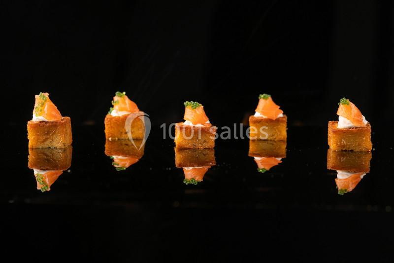 Bouchées apéritives composées de cubes dorés garnis de saumon, crème et herbes, disposés en ligne sur fond noir.