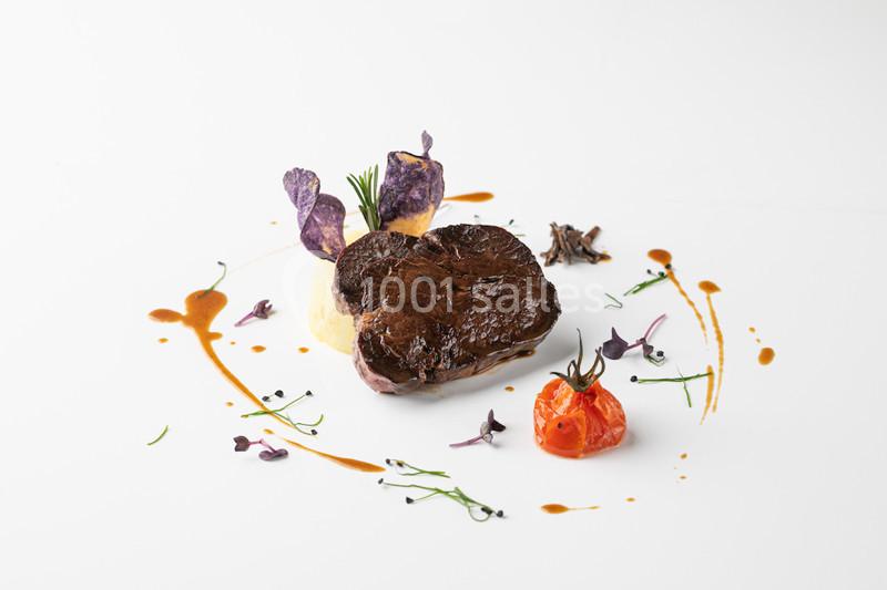 Plat gastronomique avec pièce de viande, purée, tomate confite, chips violettes et décorations de sauce et herbes.