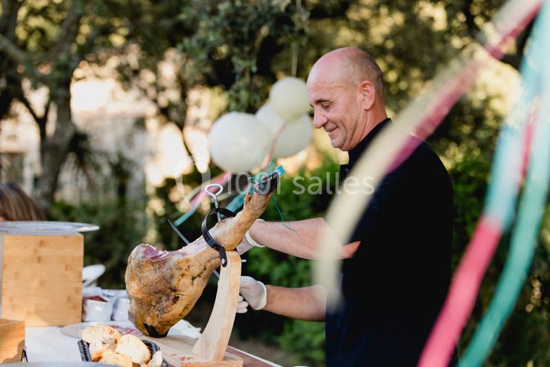 Un homme découpe un jambon sur un stand en plein air, avec des décorations et un cadre naturel en arrière-plan.