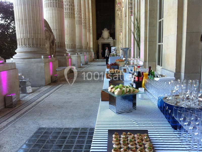 Buffet dressé sur une table rayée dans une galerie avec colonnes, comprenant amuse-bouches, verres et boissons.