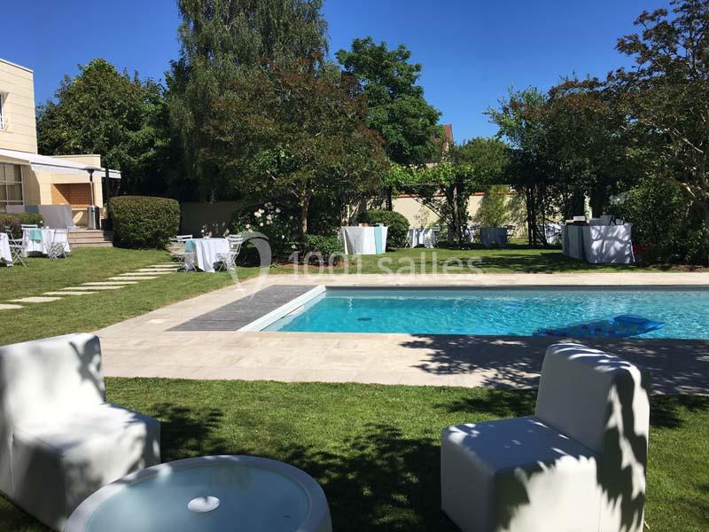 Jardin avec piscine, mobilier blanc et tables dressées, entouré de verdure sous un ciel bleu.