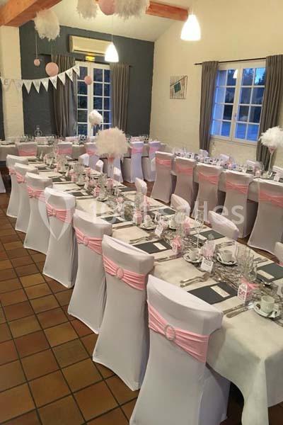 Salle décorée pour un événement avec des tables alignées, nappes blanches, chaises couvertes et rubans roses.