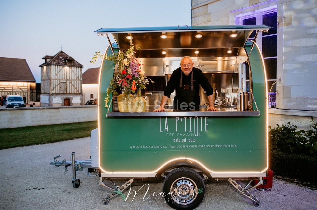 Un homme souriant se tient dans un food truck vert décoré de fleurs, stationné devant un bâtiment ancien.