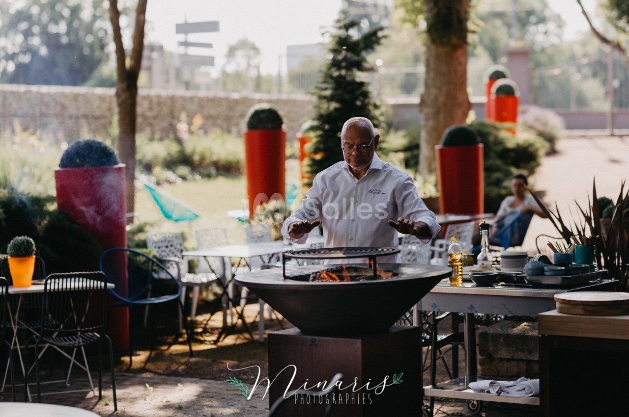 Un chef cuisine sur un brasero dans un jardin, entouré de tables et de végétation.