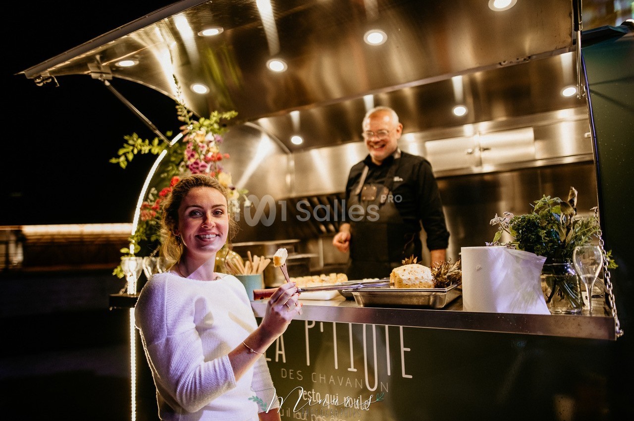 Une femme souriante tient un verre devant un food truck éclairé, avec un cuisinier en arrière-plan.