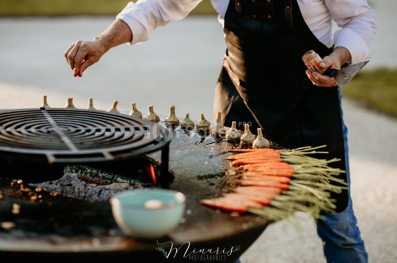 Une personne assaisonne des légumes grillés, dont des carottes et des artichauts, sur une plancha en plein air.