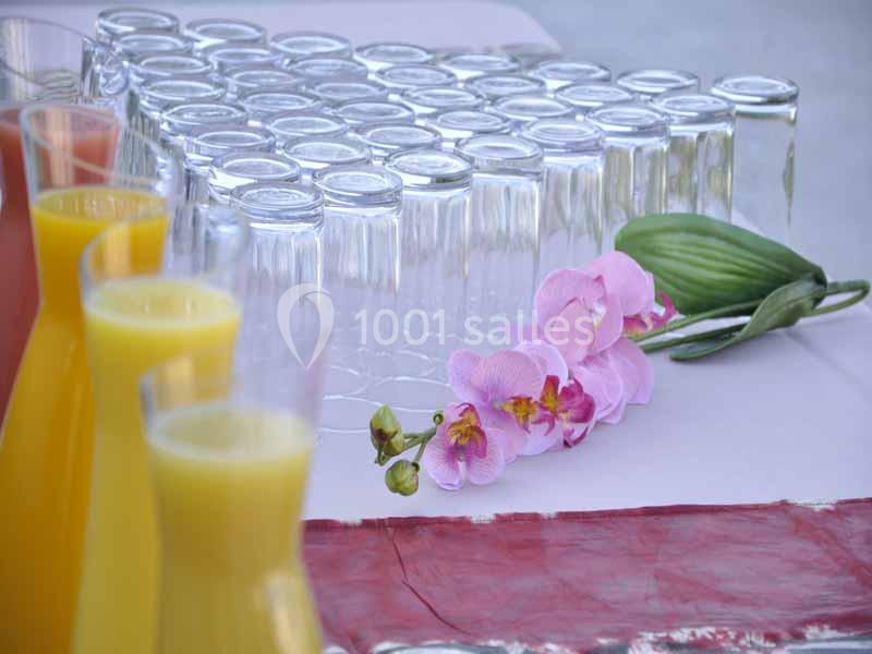 Carafes de jus colorés alignées devant des verres vides et une orchidée rose sur une nappe blanche.