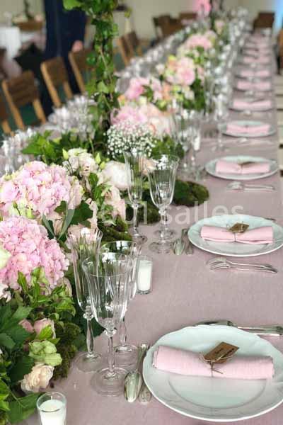 Table dressée avec une nappe rose, des assiettes blanches, des serviettes roses et une décoration florale abondante.