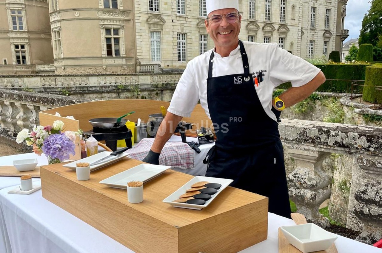 Un chef souriant en tablier noir devant une table dressée avec des plats et des ustensiles, dans un cadre extérieur élégant.