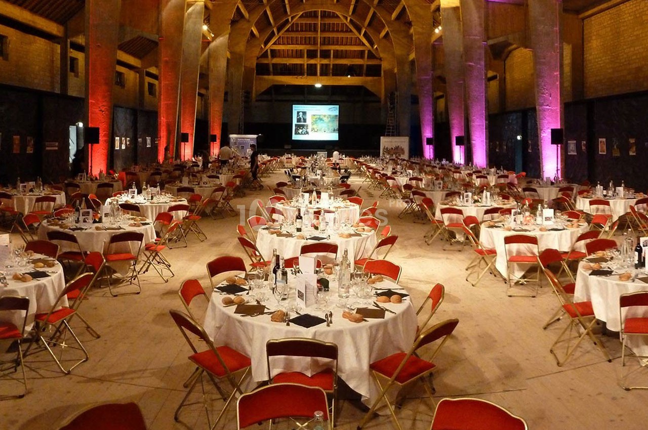 Salle spacieuse aménagée pour un dîner, avec des tables rondes dressées et des chaises rouges disposées en rangées.