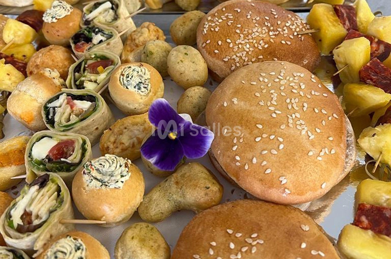 Assortiment de bouchées apéritives variées avec pains, roulés, brochettes de fruits et une fleur comestible violette.