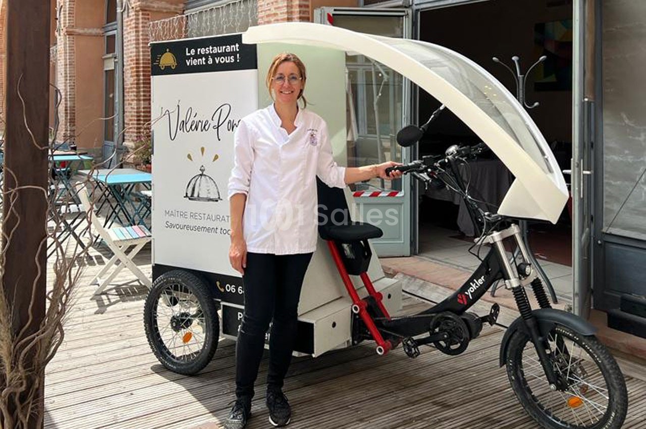 Une femme en tenue de chef pose devant un triporteur blanc stationné sur une terrasse en bois.
