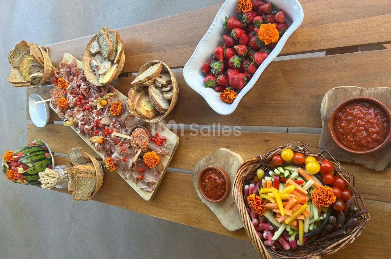 Table en bois garnie de pains, charcuteries, légumes croquants, fraises et sauces, décorée de fleurs orange.