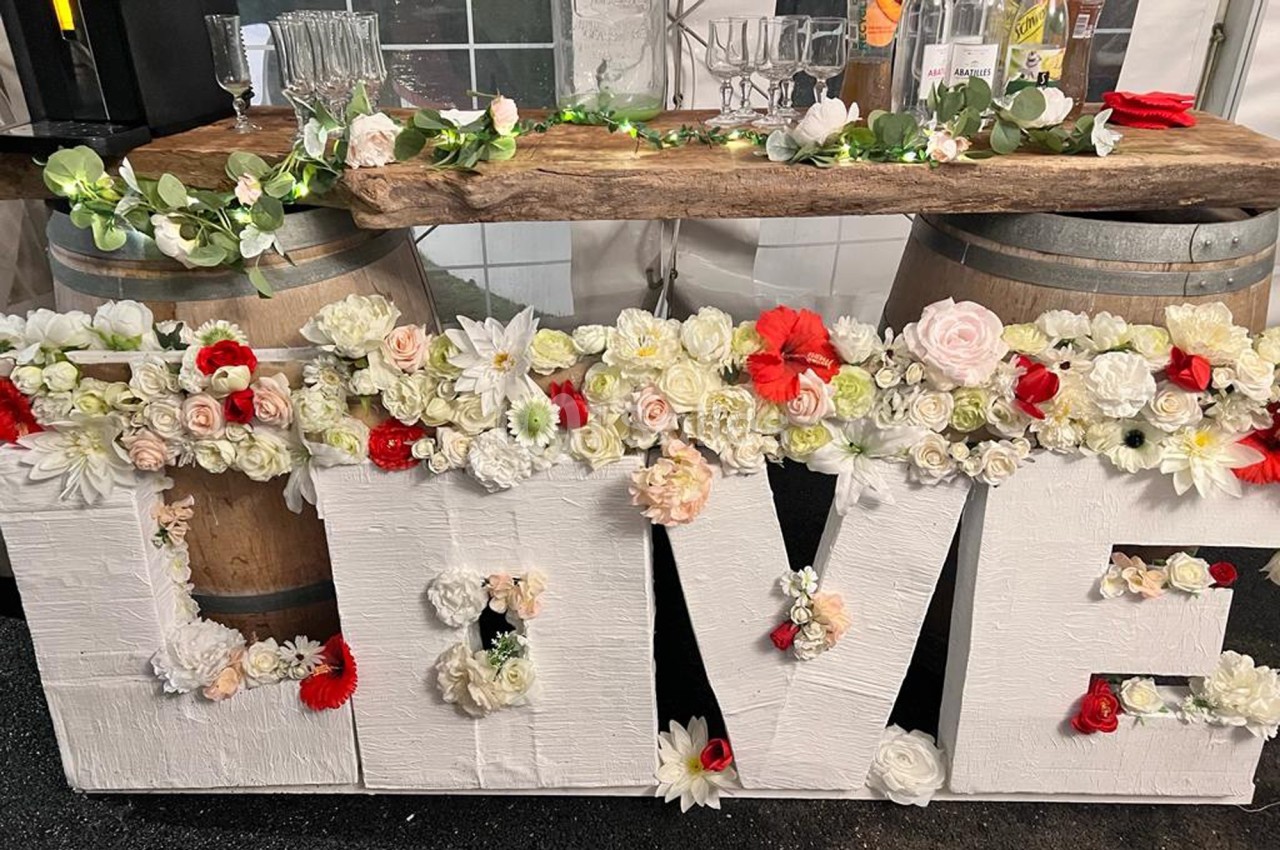 Décoration de mariage avec le mot ’LOVE’ en lettres blanches ornées de fleurs, devant une table en bois sur tonneaux.