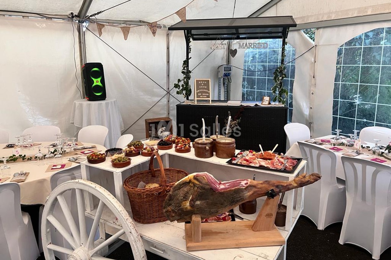 Buffet de mariage sous une tente, avec tables dressées, un jambon sur support et divers plats disposés sur une charrette.