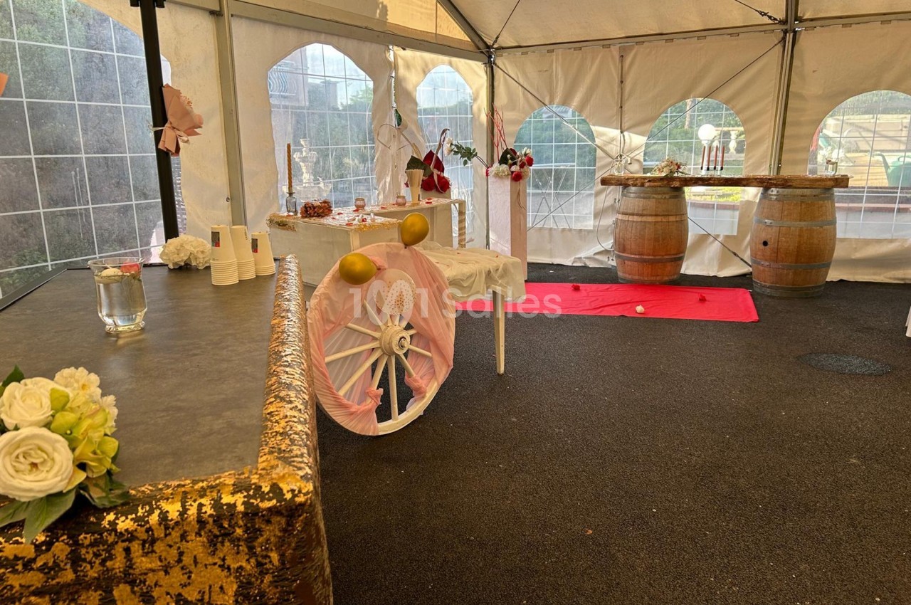 Décoration d'une tente événementielle avec tables ornées, barils en bois, fleurs et ballons dans un espace extérieur.