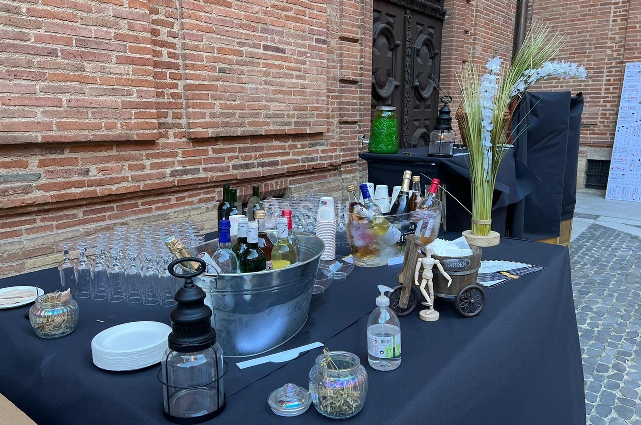 Table dressée en extérieur avec boissons, verres, seau à glace et décorations, devant un mur en briques rouges.