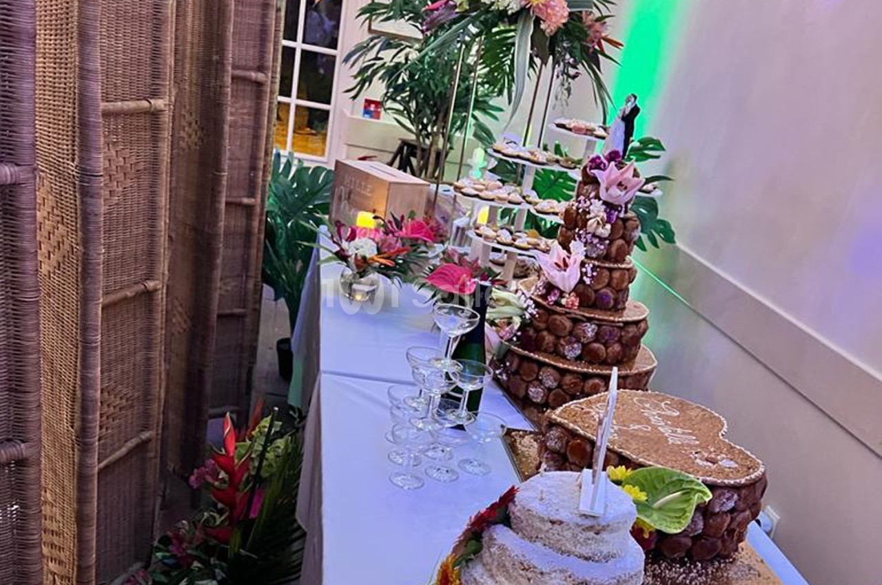 Table de desserts de fête avec pièce montée en choux, gâteau décoré, fleurs tropicales et verres empilés.