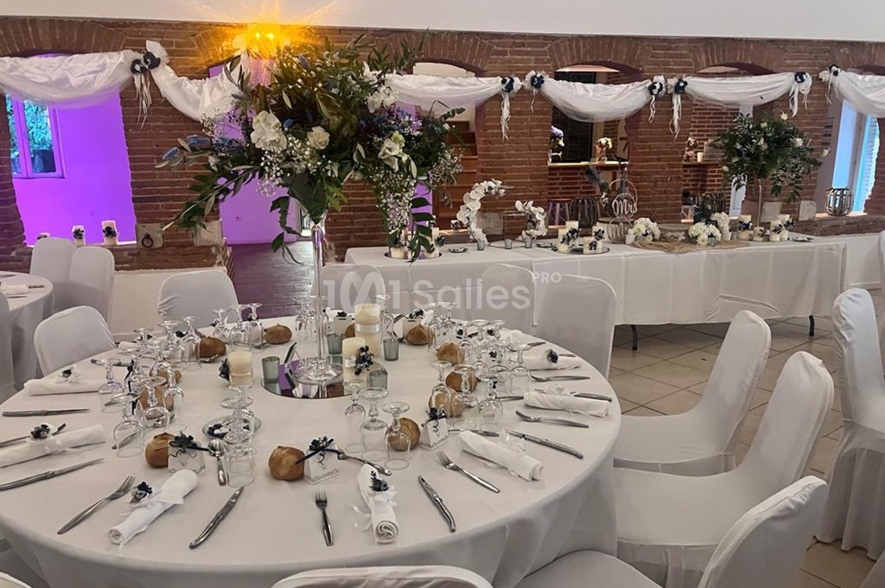 Salle de réception décorée pour un mariage avec tables rondes dressées, nappes blanches et compositions florales.