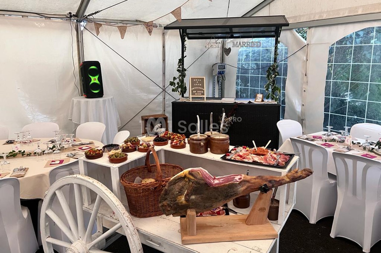 Buffet de mariage sous une tente, avec tables dressées, un jambon sur support et divers plats disposés sur une charrette.