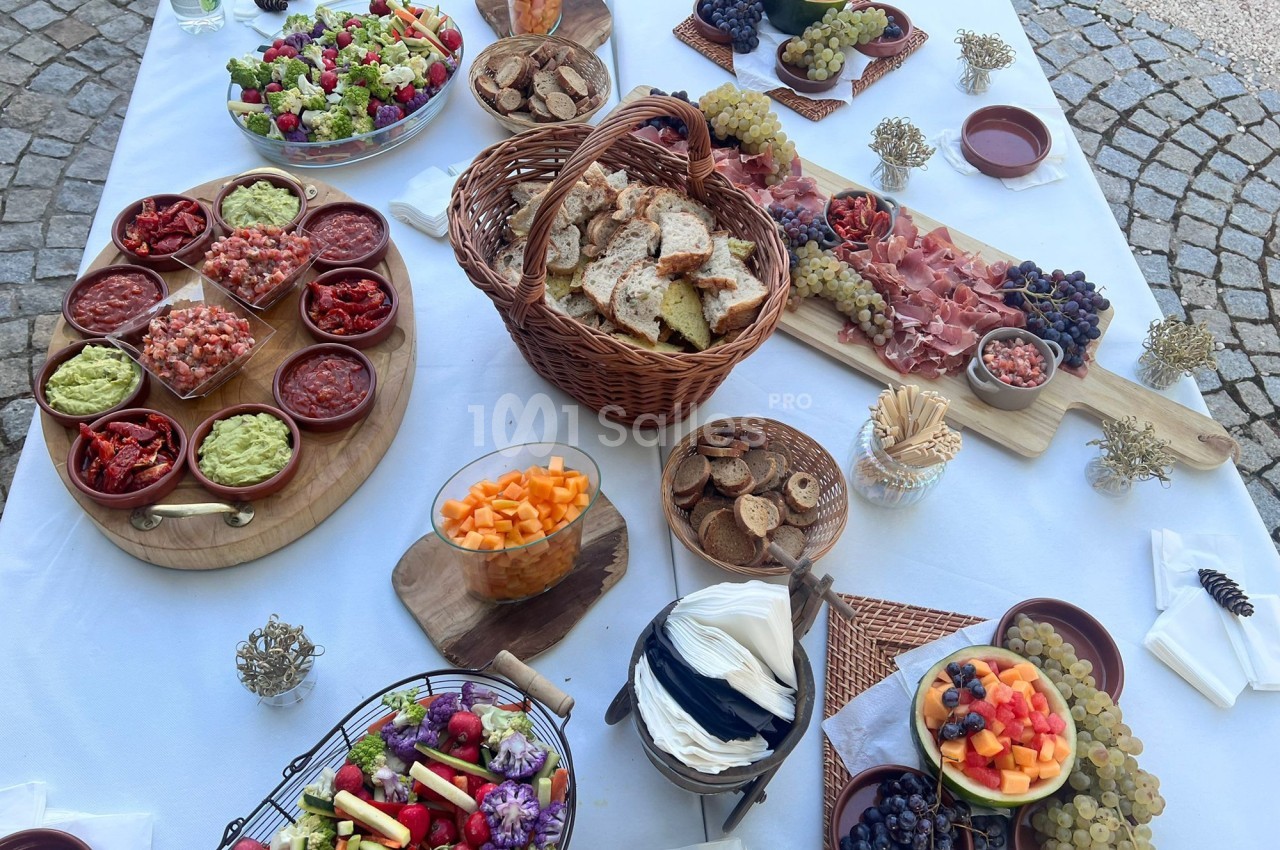 Table garnie de plats variés : salades, fruits, charcuterie, pain, dips et fromages, disposés sur une nappe blanche.