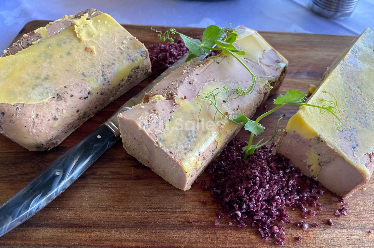Bloc de foie gras tranché sur une planche en bois, accompagné de sel violet et décoré de pousses vertes.