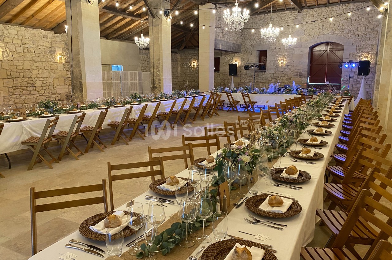 Grande salle en pierre décorée pour un événement, avec longues tables dressées, chaises en bois et éclairage chaleureux.