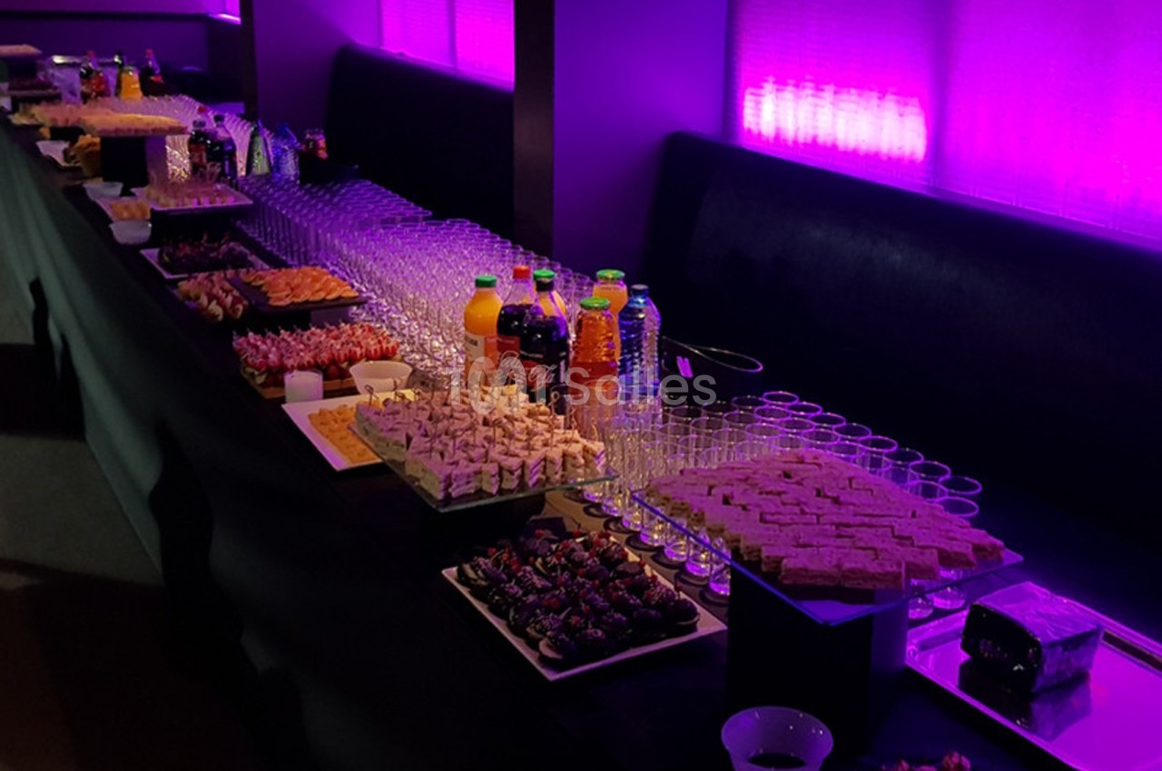 Buffet avec assortiments de boissons, verrines, canapés et desserts, disposé sur une table éclairée par une lumière violette.