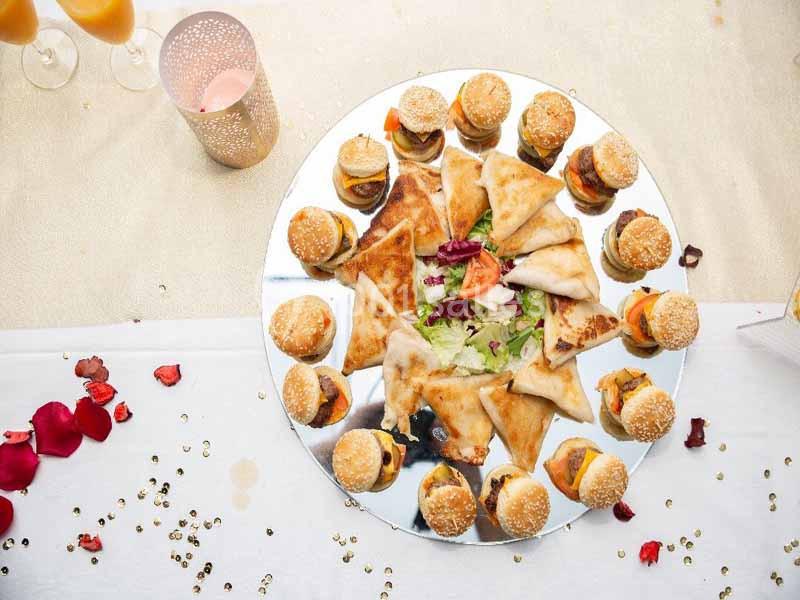 Plateau de mini-burgers et samoussas disposés autour d'une salade, sur une table décorée de pétales et paillettes.