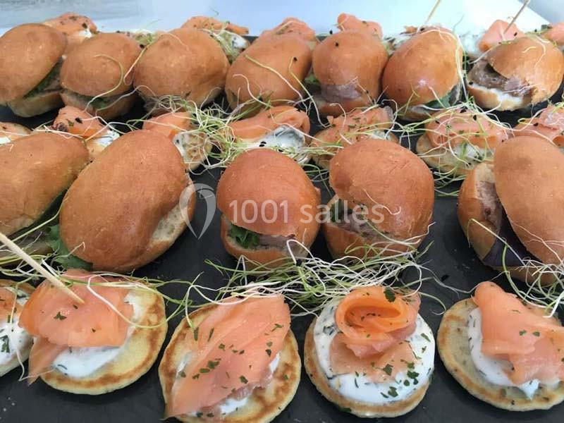 Assortiment de mini-burgers et blinis garnis de saumon fumé, crème et pousses vertes, présentés sur un plateau.
